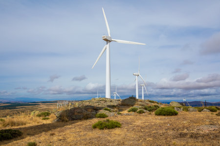 Several wind turbines rise above a grassy landscape with rocky outcrops, showcasing sustainable energy production under a bright, partially cloudy sky.の写真素材