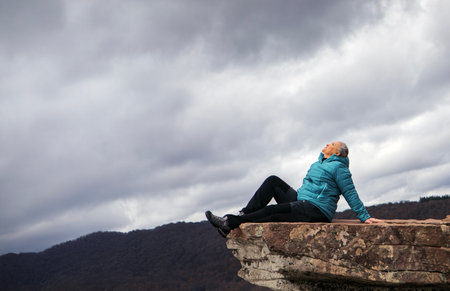 A woman relaxes on the edge of a rocky cliff, gazing upwards into the dark, swirling clouds overhead. Surrounding mountains create a serene backdrop, highlighting her connection with nature.の写真素材