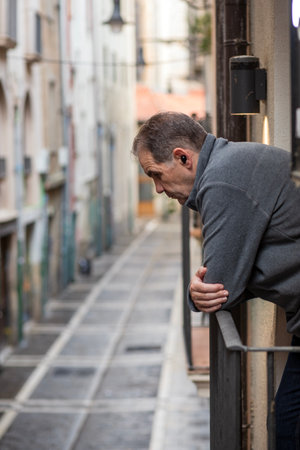 A man leans on a balcony, reflecting deeply as he looks down a narrow, empty street lined with charming buildings. The scene captures the calm ambiance of an early day.の写真素材