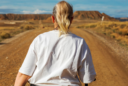 A woman with long hair, wearing a simple white shirt, strolls down a gravel road. The scene captures the beauty of an expansive desert landscape, featuring rugged cliffs and natural serenity.の写真素材