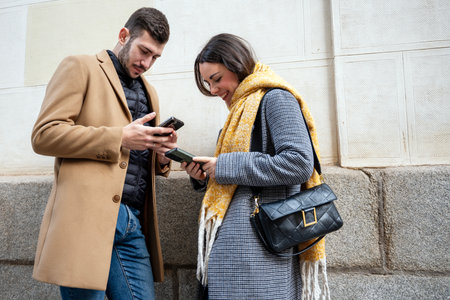 Two young adults stand close on a bustling street, engaged with their mobile devices. They smile, sharing a moment as they explore their screens while wrapped in stylish winter outfits.の写真素材