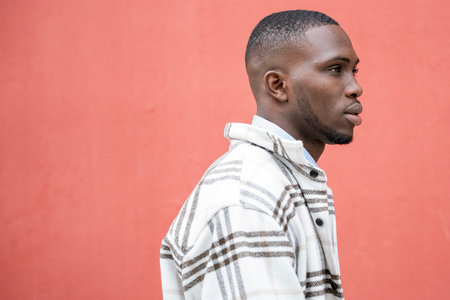 A young man stands with poise along a bold red wall, wearing a trendy plaid shirt. The urban...