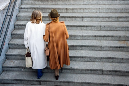 Two women stroll down a broad set of stairs, clad in fashionable coats. One wears a white coat and the other a tan wrap. Their casual conversation brightens the vibrant atmosphere of the city plaza.の写真素材