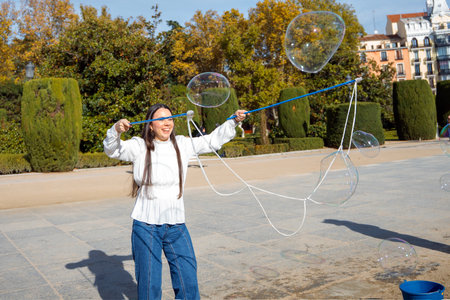 A young person delights in creating large bubbles using long strings in a beautiful park. The sun...