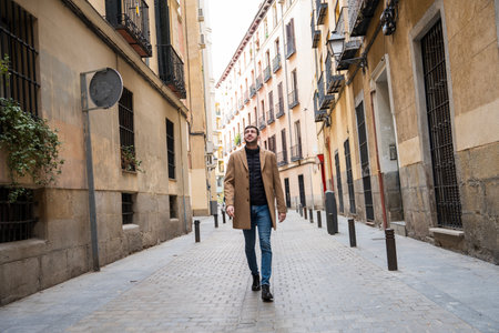 A man strolls confidently down a narrow street in Madrid, surrounded by historic buildings. Sunlight filters through, creating a warm atmosphere in this vibrant neighborhood.の写真素材