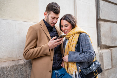 A young couple stands close together on a city street, sharing a smile as they look at a smartphone....