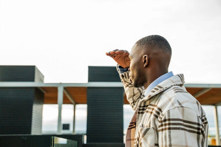 A man stands on a rooftop, shielding his eyes from the sun as he gazes into the distance. With a stylish coat and confident posture, he enjoys the view of a bustling cityscape below.の写真素材