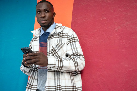 A young man leans against a vibrant wall splashed with colors while looking at his smartphone. The...