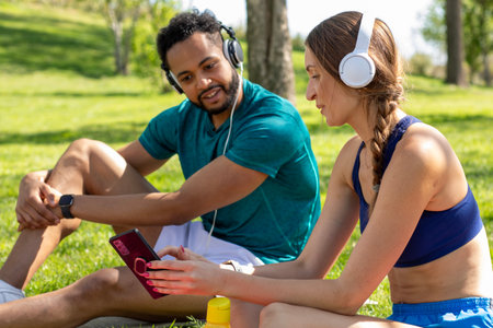 Two friends sit on the grass in a lush park, sharing music through headphones. The warm sun shines...