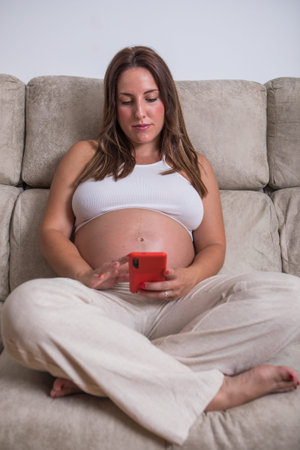 A pregnant woman sits comfortably on a soft couch, dressed in casual clothes. She holds her phone, engaging with family or friends, glowing with happiness and anticipation for her new arrival.の写真素材