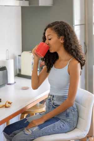 A young woman sits comfortably at a kitchen counter, sipping from a bright mug. She wears a casual tank top and jeans, enjoying a moment of peace with a pastry nearby.の写真素材