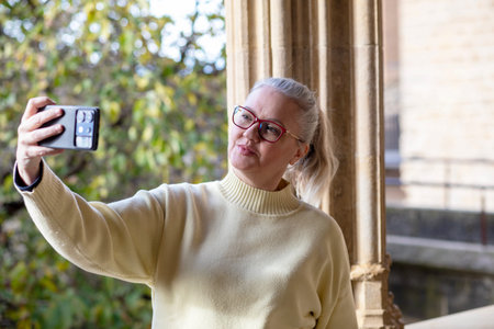 A woman with glasses takes a selfie against a backdrop of beautiful architecture and greenery. She...