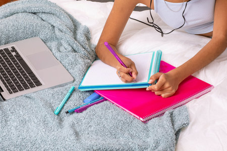 A student sits comfortably in bed, writing notes in vibrant notebooks while listening to music. A laptop rests nearby, creating a productive and cozy atmosphere for studying.の写真素材