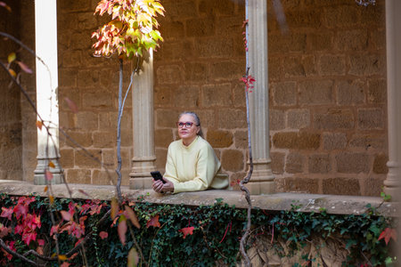 A person sits in a garden space, surrounded by vibrant autumn foliage, lost in thought while holding a phone, enjoying the peaceful atmosphere.の写真素材