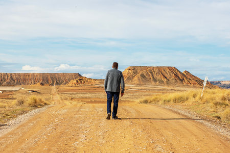 A solitary figure walks down a quiet dirt road, embraced by vast red rock cliffs and an expansive sky. The serene atmosphere invites reflection and tranquility amid natures beauty.の写真素材