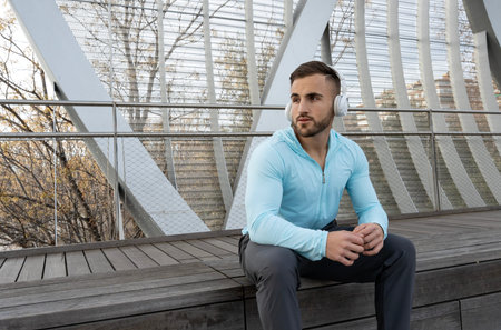 A young man sits quietly on a wooden deck, wearing a light blue hoodie and headphones. Surrounding him are minimalist architectural elements and sparse trees, creating a serene atmosphere.の写真素材