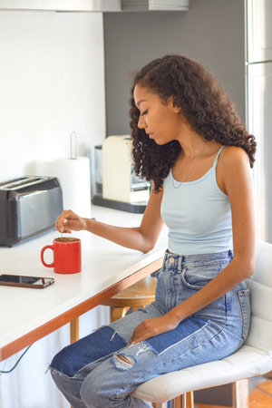 A young woman sits at a modern kitchen counter, stirring her coffee as sunlight spills through the window. She wears a light blue top and denim jeans, creating a relaxed vibe.の写真素材