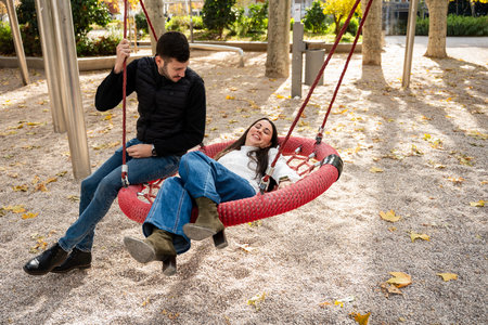 Two friends share laughter while swinging side by side in a colorful park filled with autumn leaves....