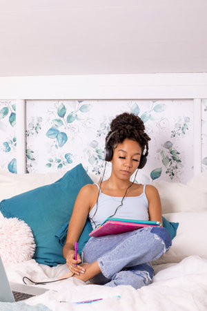 In a serene bedroom filled with soft colors, a young woman enjoys studying. She is focused on her notebook, listening to music through headphones, surrounded by cushions.の写真素材