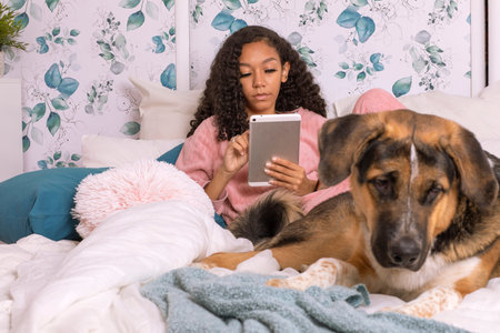 A girl sits comfortably on a bed, focused on her tablet as her dog lies peacefully next to her. The room features soft colors and a serene atmosphere, perfect for relaxation.の写真素材