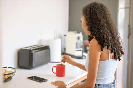 A woman with curly hair enjoys her cozy kitchen. She stirs her coffee in a bright red mug while morning sunlight bathes the space, creating a lovely atmosphere.の写真素材
