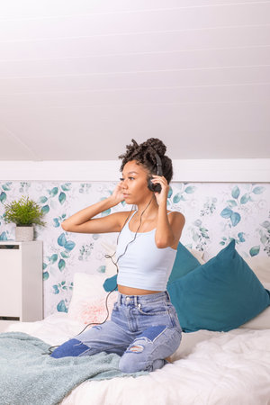 A young woman sits on a bed, lost in music while wearing headphones. Soft floral decor and cozy cushions create a relaxing atmosphere, perfect for unwinding.の写真素材