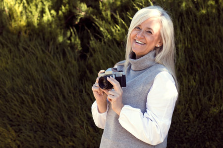 A cheerful woman in her 50s stands outdoors, holding a vintage camera. Dressed in a cozy grey vest...