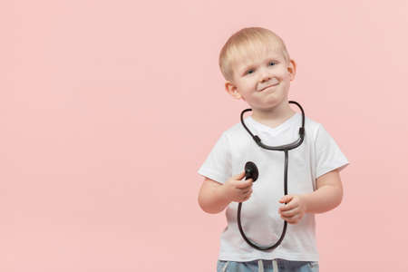 Child blond boy stands and holds a stethoscope or phonendoscope in his hands on a pink background with place for text.の写真素材