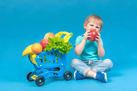 A hungry blond child sits near a supermarket basket full of food carts and bites a huge red apple. The concept of food delivery and healthy eating fruits and vegetables. Studio shot for advertising.の写真素材