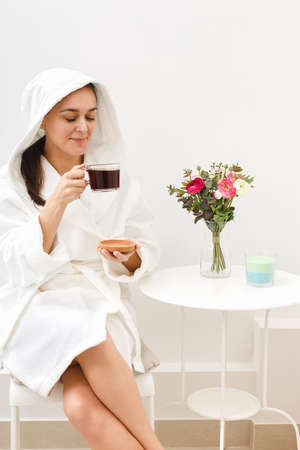 A young girl drinks coffee in a relaxation room in a beauty salon after beauty treatments or a spa sauna. The concept of care and customer-oriented services in medical center. Vertical studio shot.の写真素材