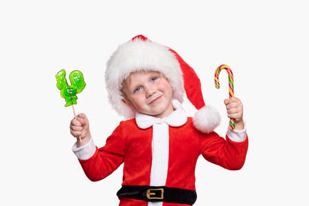 A child boy 4 years old in a New Year costume and a hat of Santa Claus holds candies in his hands. Cheerful positive joyful concept about holidays and gifts. Studio shot on white background isolate.の写真素材