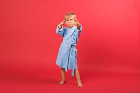 A blond child boy in blue bathrobe on a red bright background shows a hand sign shoots gun or aims target near eyes. Studio shot of a child dancing in bathhouse, sauna, spa, pool and swimming school.の写真素材