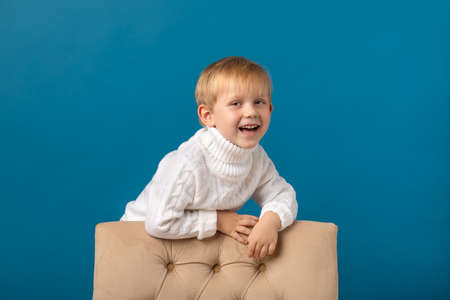 A joyful boy, a blond child in a white warm knitted sweater, stands near the back of a chair, looks at the camera and laughs. Winter studio shot on a bright blue background.の写真素材