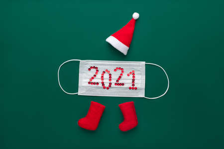 Concept for new year 2021 and christmas. Red santa claus hat and felt boots on green background with medical protective mask from coronavirus covid-19. The festive text is lined with shiny sequins.の写真素材