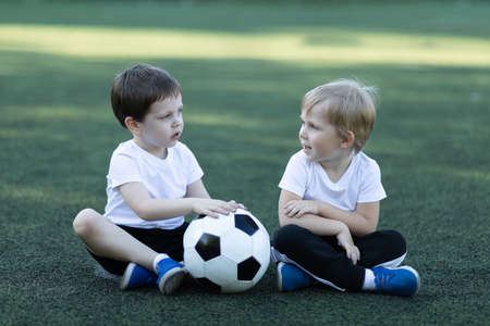 Lifestyle of two children and soccer ball, sitting on grass, laughing and talking. Concept about childhood, sports and communication of preschool boys.の写真素材