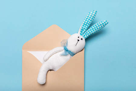 White toy hare sewn from fabric in paper envelope with letter or postcard. Studio shot of flat lay,...