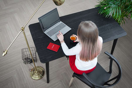 Top view, young woman sitting at table and working on computer laptop, drinking tea. Office work concept.の写真素材