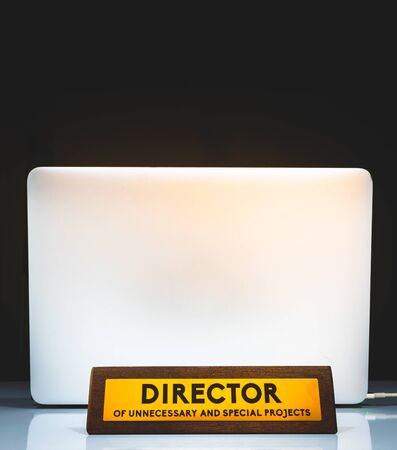 Office funny plate with title : "Director of unnecessary and special projects" with silver laptop on the background and black backround. Concept of absence at work.の写真素材