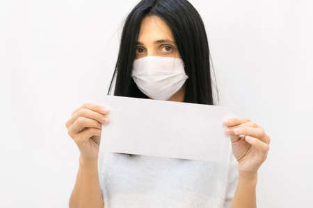 Close up of young caucasian woman with white mask on holding white envelope with blank space and white wall. Conceptual note background.の写真素材