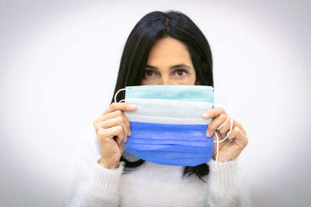 Brunette woman holds three surgical face masks close to face covering mouth isolated in grey background. Blue; green and white mask. Fashion and variety of corona accessories.の写真素材