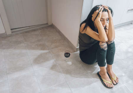 Young attractive  caucasian woman  sits on the kitchen florr tired with hands on her head and full glass of wine.の写真素材