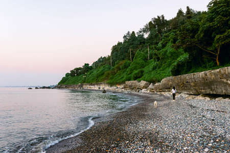 Woman with stray dog walks on rocky beach on the coastline of Black sea with calm sea and green nature in the background. Tiskhisdziri village in Adjara.の写真素材