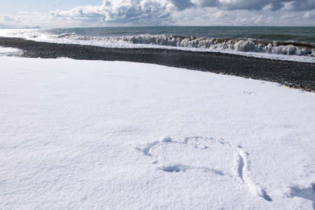 Heart shaped form on white snow with rocky beacj and sea view onthe background. Horizontal blank space image.の写真素材