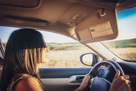Young attractive girl in drivers seat looks straight and holds hands on steering wheel with nature outside the window in the backgroundの写真素材
