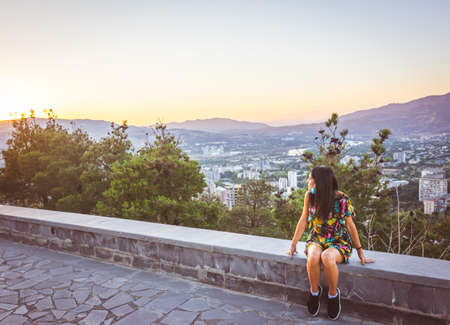 Young girl sits isolated with mask off and looks to the sunset with panoramic view of the city in...