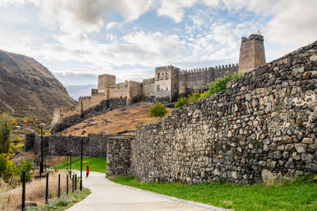 Female tourist stands on the path next to huge Khertvisi fortress wall with towers above in cloudy day. Travel copyspace background.のeditorial素材