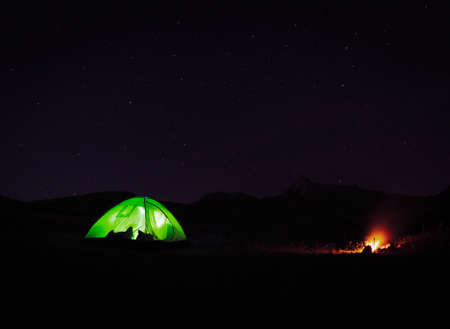 Illuminated Green tent standing by camp fire in nature at night with starry night background.