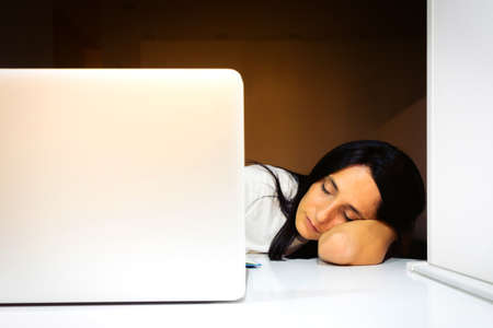 Young beautiful woman sleeping face on the elbow with  laptop nearby on white desktopの写真素材