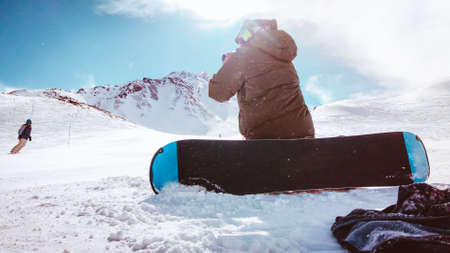 Photographer snowboarder on his knees on position on slope shooting skier skiing downhill in mountains ski resort in winterの写真素材