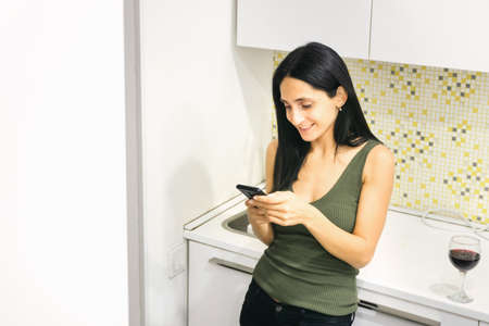 Young caucasian woman holds a smartphone with smiley face and wine glass on kitchen topの写真素材